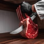 A butcher wearing food safety gloves and cutting through a raw tomahawk steak