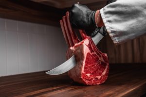 A butcher wearing food safety gloves and cutting through a raw tomahawk steak